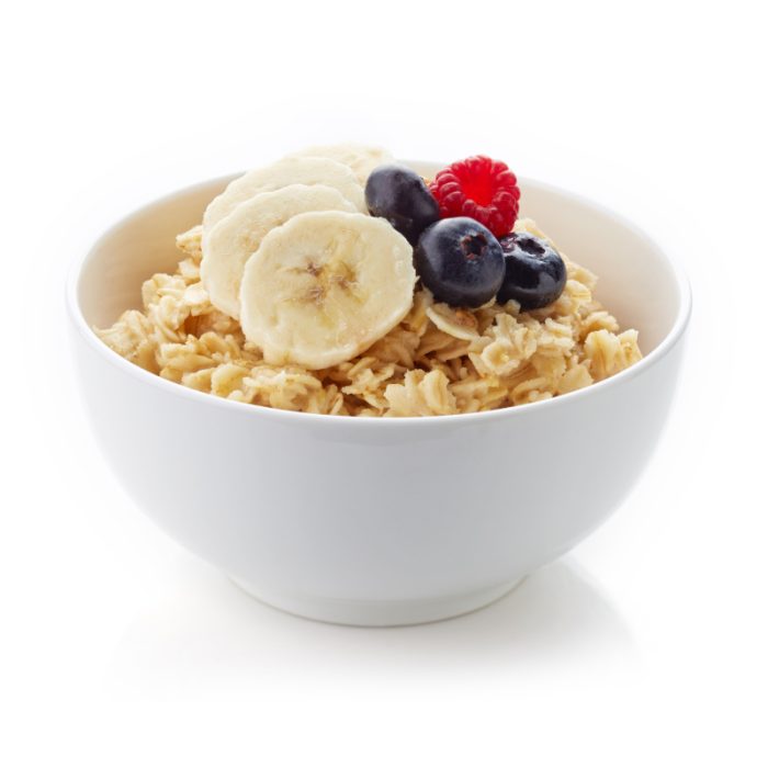 Tazón de cereales con plátano, arándanos y frambuesas sobre fondo blanco.